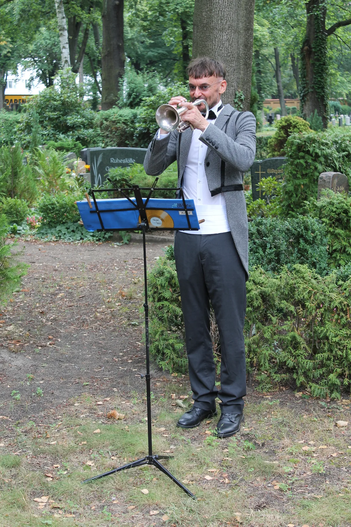 Ron Dietze spielt Trompete auf einem Friedhof unter alten Bäumen